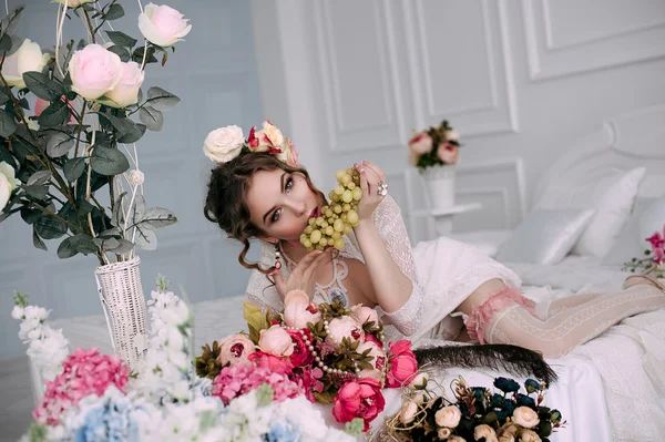 Beautiful young sexy woman sitting on white bed and eating grapes, wearing white lace dress, room decorated with flowers. Perfect makeup. Beauty fashion. Eyelashes. Studio retouched shot.