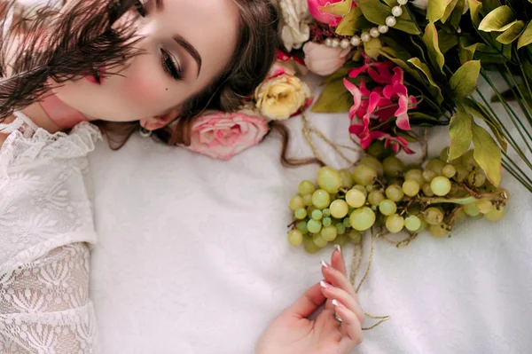 Beautiful young sexy woman sitting on white bed and eating grapes, wearing white lace dress, room decorated with flowers. Perfect makeup. Beauty fashion. Eyelashes. Studio retouched shot.