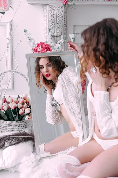 Beautiful young sexy woman , looking in the mirror wearing white lingerie, room decorated with flowers. Perfect makeup. Beauty fashion. Eyelashes. Studio retouched shot.