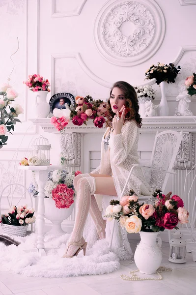 Beautiful young sexy woman , holding a glass and sitting in a chair, wearing white lingerie, room decorated with flowers. Perfect makeup. Beauty fashion. Eyelashes. Studio retouched shot.