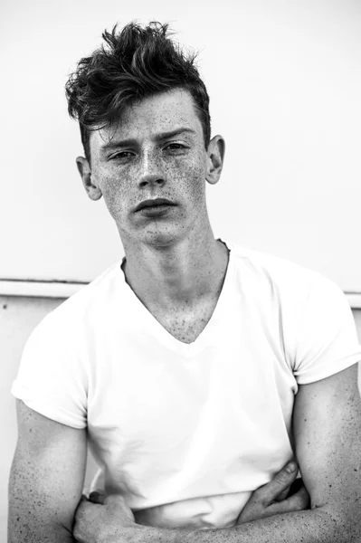 Portrait of attractive stylish young guy model with red hair and freckles sitting on green grass, wearing jacket. Fashionable outdoor shot. Black and white image, close up