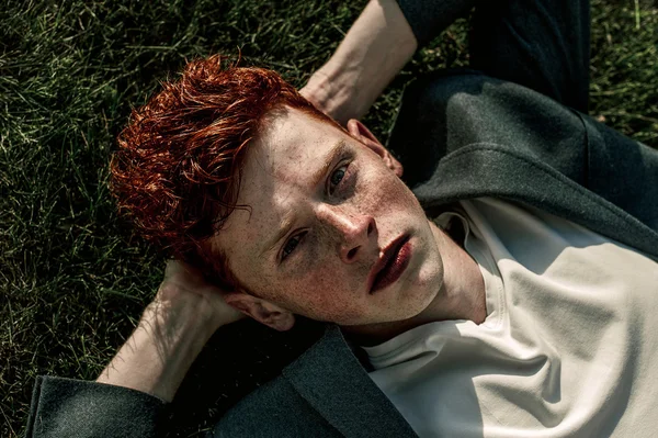 Portrait of attractive stylish young guy model with red hair and freckles sitting on green grass, wearing jacket. Fashionable outdoor shot. Close up
