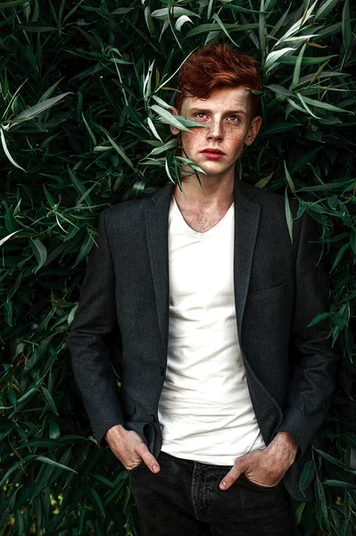 Portrait of attractive stylish young guy model with red hair and freckles sitting on green grass, wearing jacket. Fashionable outdoor shot