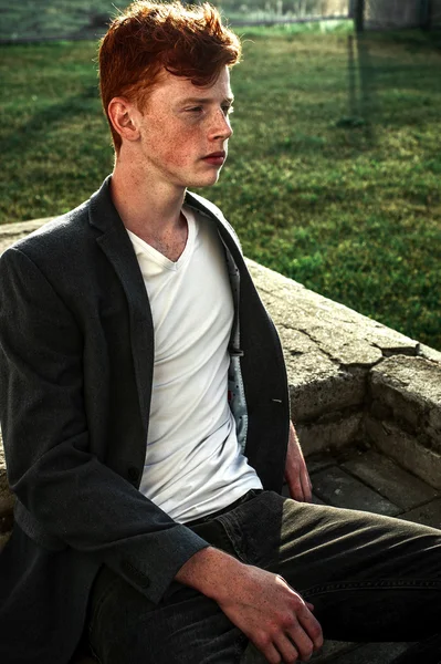 Portrait of attractive stylish young guy model with red hair and freckles sitting on green grass, wearing jacket. Fashionable outdoor shot