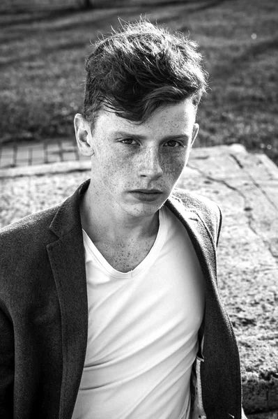 Portrait of attractive stylish young guy model with red hair and freckles sitting on green grass, wearing jacket. Fashionable outdoor shot. Black and white image