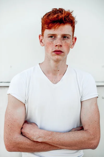 Portrait of attractive stylish young guy model with red hair and freckles standing near white wall , wearing white t-shirt. Fashionable outdoor shot.