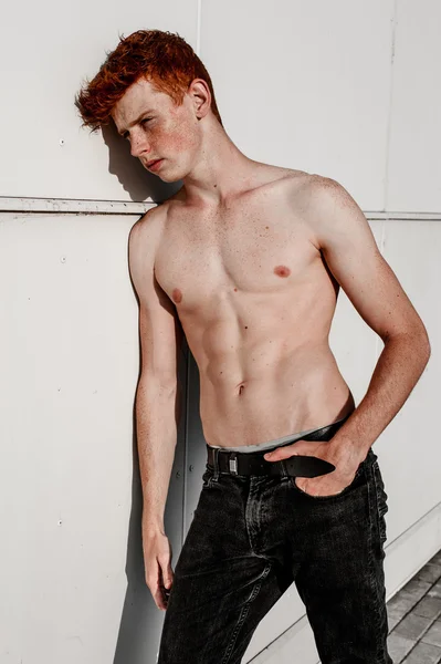 Portrait of attractive stylish young guy model with red hair and freckles standing near white wall , without t-shirt. Fashionable outdoor shot.