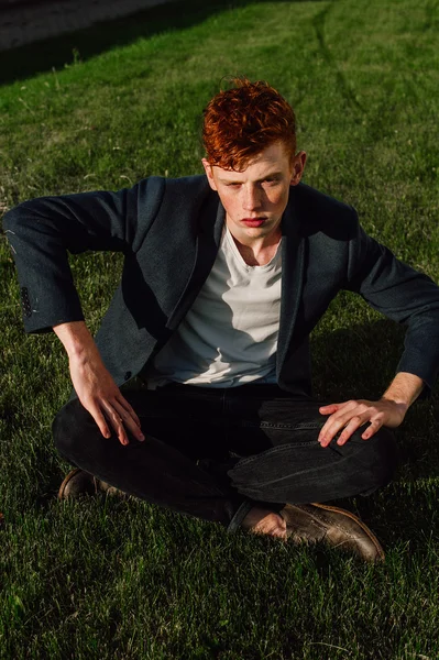 Portrait of attractive stylish young guy model with red hair and freckles sitting on green grass, wearing jacket. Fashionable outdoor shot