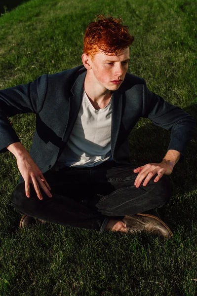 Portrait of attractive stylish young guy model with red hair and freckles sitting on green grass, wearing jacket. Fashionable outdoor shot