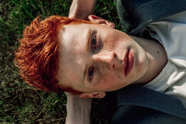 Portrait of attractive stylish young guy model with red hair and freckles sitting on green grass, wearing jacket. Fashionable outdoor shot