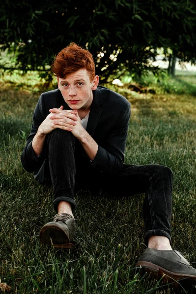 Portrait of attractive stylish young guy model with red hair and freckles sitting on green grass, wearing jacket. Fashionable outdoor shot