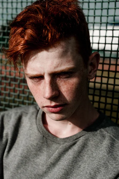 One young handsome red haired fashionable boy standing near stadium in sunny day outdoor , wearing casual clothing