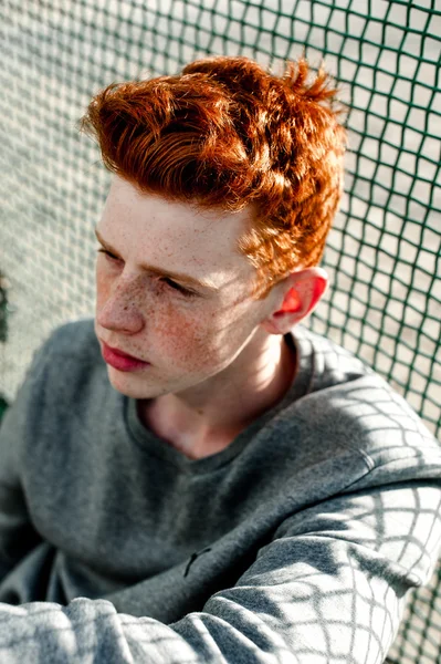 One young handsome red haired fashionable boy standing near stadium in sunny day outdoor , wearing casual clothing