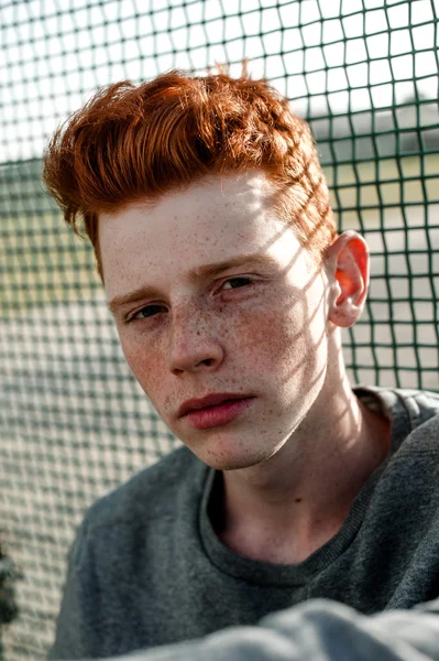 One young handsome red haired fashionable boy standing near stadium in sunny day outdoor , wearing casual clothing