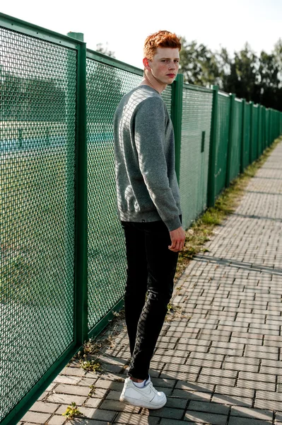 One young handsome red haired fashionable boy standing near stadium in sunny day outdoor , wearing casual clothing