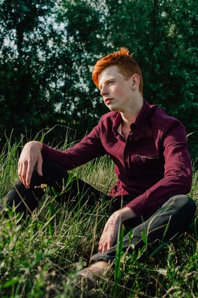 Portrait of attractive stylish young guy model with red hair and freckles sitting on green grass, wearing purple shirt. Fashionable outdoor shot