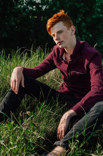 Portrait of attractive stylish young guy model with red hair and freckles sitting on green grass, wearing purple shirt. Fashionable outdoor shot