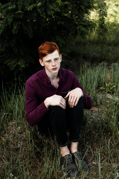 Portrait of attractive stylish young guy model with red hair and freckles sitting on green grass, wearing purple shirt. Fashionable outdoor shot