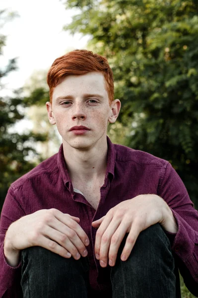Portrait of attractive stylish young guy model with red hair and freckles sitting on green grass, wearing purple shirt. Fashionable outdoor shot