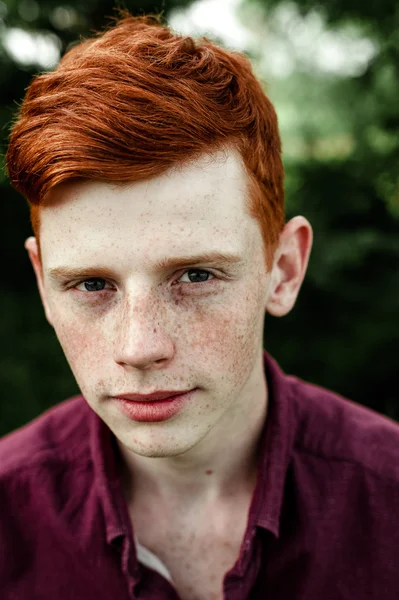 Portrait of attractive stylish young guy model with red hair and freckles sitting on green grass, wearing purple shirt. Fashionable outdoor shot. Close up