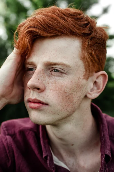 Portrait of attractive stylish young guy model with red hair and freckles sitting on green grass, wearing purple shirt. Fashionable outdoor shot. Close up