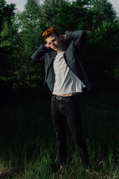 Portrait of attractive stylish young guy model with red hair and freckles standing on green grass, wearing jacket. Fashionable outdoor shot.