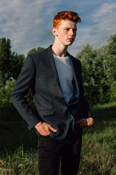 Portrait of attractive stylish young guy model with red hair and freckles standing on green grass, wearing jacket. Fashionable outdoor shot.