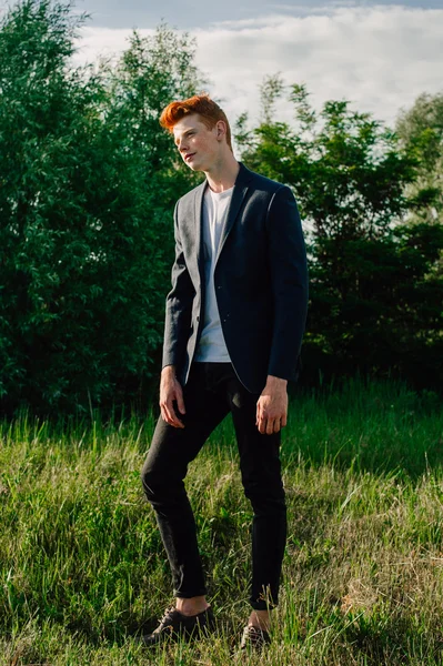 Portrait of attractive stylish young guy model with red hair and freckles standing on green grass, wearing jacket. Fashionable outdoor shot.