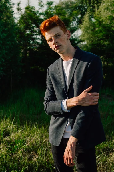 Portrait of attractive stylish young guy model with red hair and freckles standing on green grass, wearing jacket. Fashionable outdoor shot.