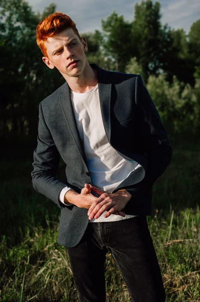 Portrait of attractive stylish young guy model with red hair and freckles standing on green grass, wearing jacket. Fashionable outdoor shot.