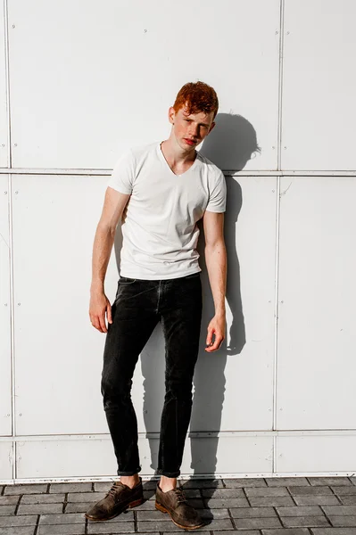 Portrait of attractive stylish young guy model with red hair and freckles standing near white wall , wearing white t-shirt. Fashionable outdoor shot.