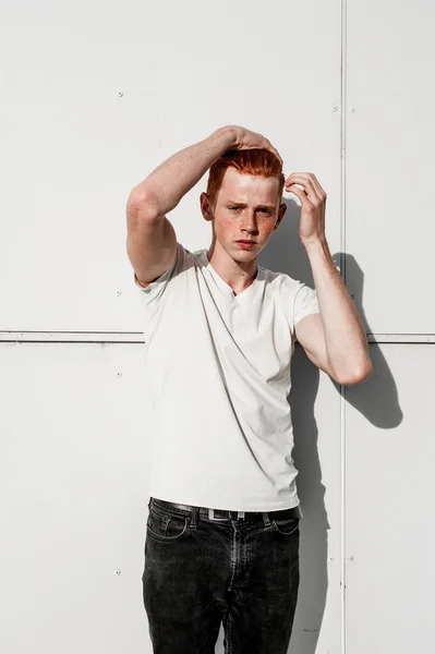 Portrait of attractive stylish young guy model with red hair and freckles standing near white wall , wearing white t-shirt. Fashionable outdoor shot.