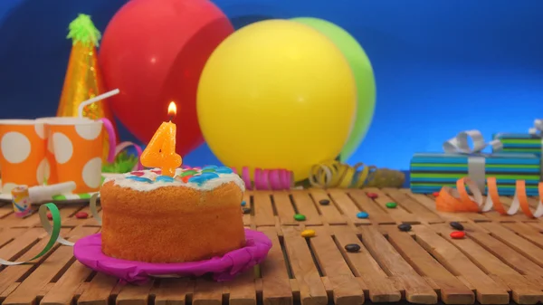 Birthday cake with candles on rustic wooden table with background of colorful balloons, gifts, plastic cups and candies with blue wall in the background