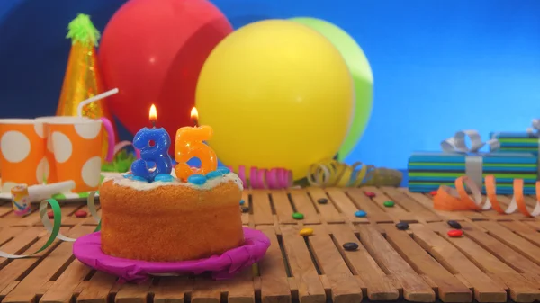 Birthday cake with candles on rustic wooden table with background of colorful balloons, gifts, plastic cups and candies with blue wall in the background