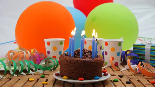 Chocolate birthday cake with a blue candles burning on rustic wooden table with background of colorful balloons, gifts, plastic cups with candies and white wall in the background