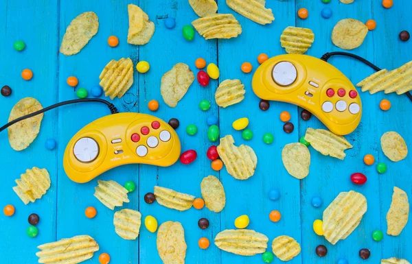 Two Game controller, chips and candy on a blue wooden background. fast food, snacks, not unhealthy food. Colored candy. Snack between games. Colored pills. Video game console GamePad.