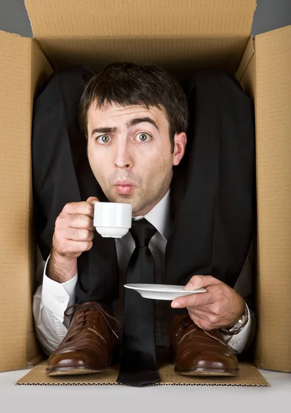 Flexible businessman with doubtful face in a cardboard box drinking coffee