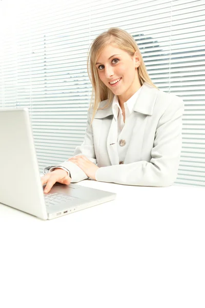 Smiling blond businesswoman with laptop near venetian blind window