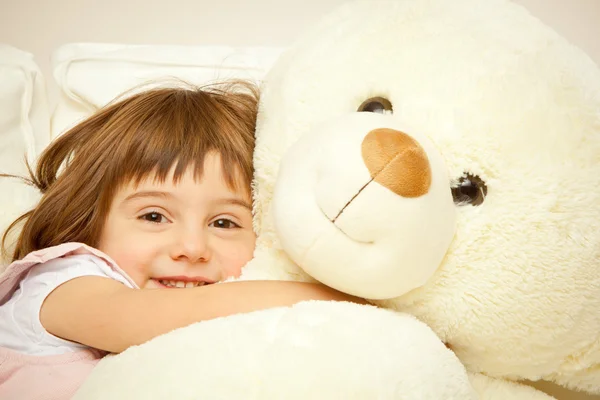 Blonde female child play with her white teddy bear in a bed