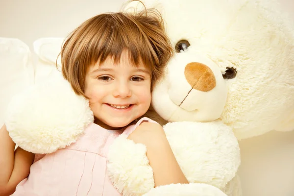 Blonde female child play with her white teddy bear in a bed