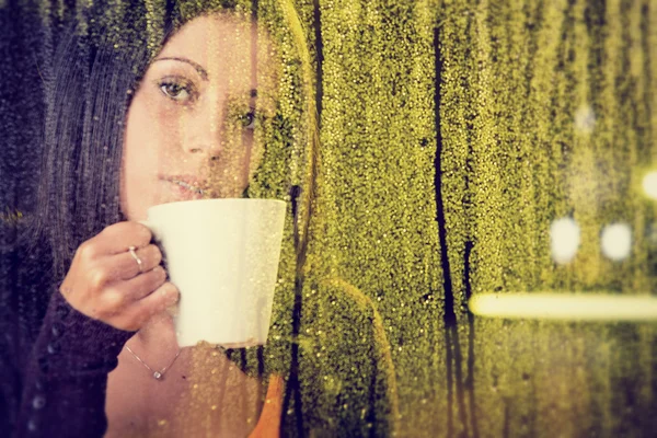 Romantic young woman with cup of tea looks at rain through the window