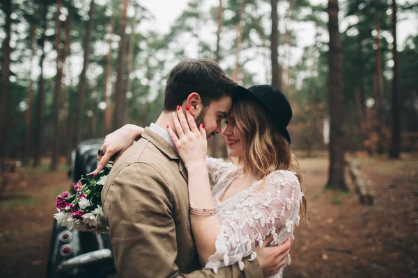 Stylish Loving wedding couple kissing and hugging in a pine forest near retro car