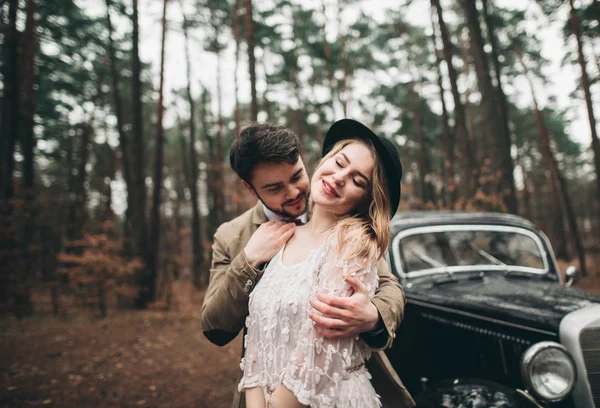 Stylish Loving wedding couple kissing and hugging in a pine forest near retro car