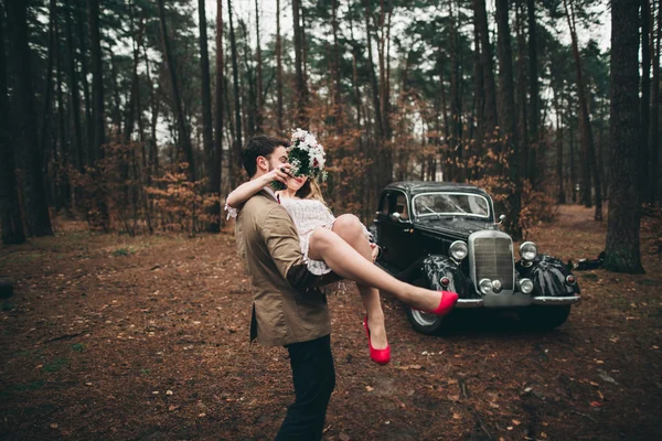 Gorgeous newlywed bride and groom posing in pine forest near retro car in their wedding day