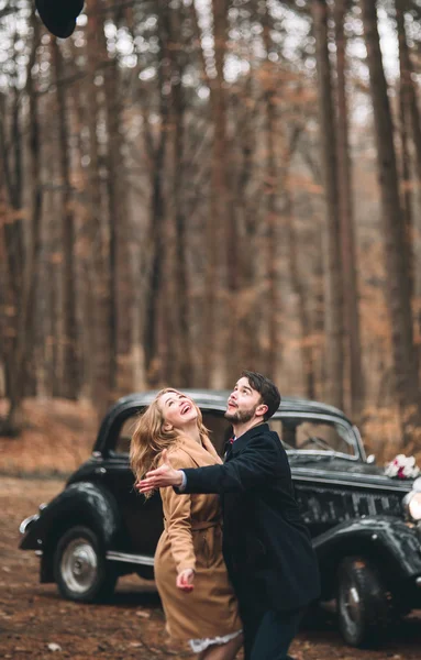 Gorgeous newlywed bride and groom posing in pine forest near retro car in their wedding day