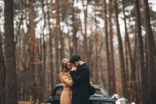 Gorgeous newlywed bride and groom posing in pine forest near retro car in their wedding day