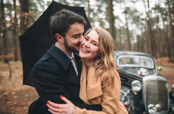 Gorgeous newlywed bride and groom posing in pine forest near retro car in their wedding day