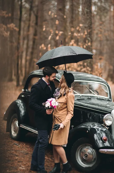 Stylish Loving wedding couple kissing and hugging in a pine forest near retro car