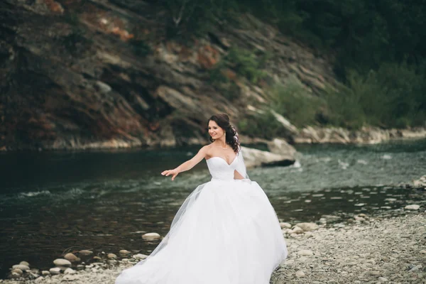 Beautiful luxury young bride in long white wedding dress and veil standing near river with mountains on background