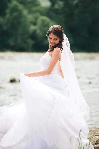 Beautiful luxury young bride in long white wedding dress and veil standing near river with mountains on background